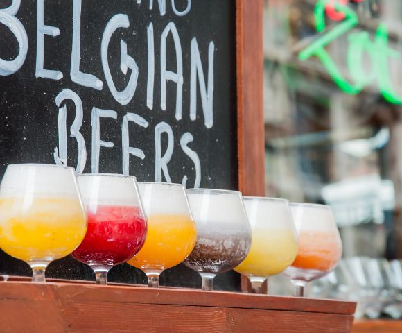 Colorful Belgian beers in tasting glasses line a bar beneath a chalkboard reading “Belgian Beers,” creating a lively, inviting pub atmosphere.