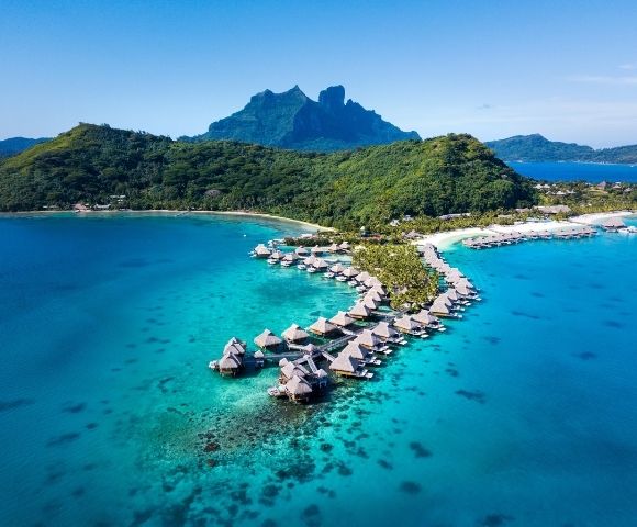 Aerial view of a tropical resort with thatched-roof overwater bungalows extending into a turquoise lagoon, with lush green mountains in the background.