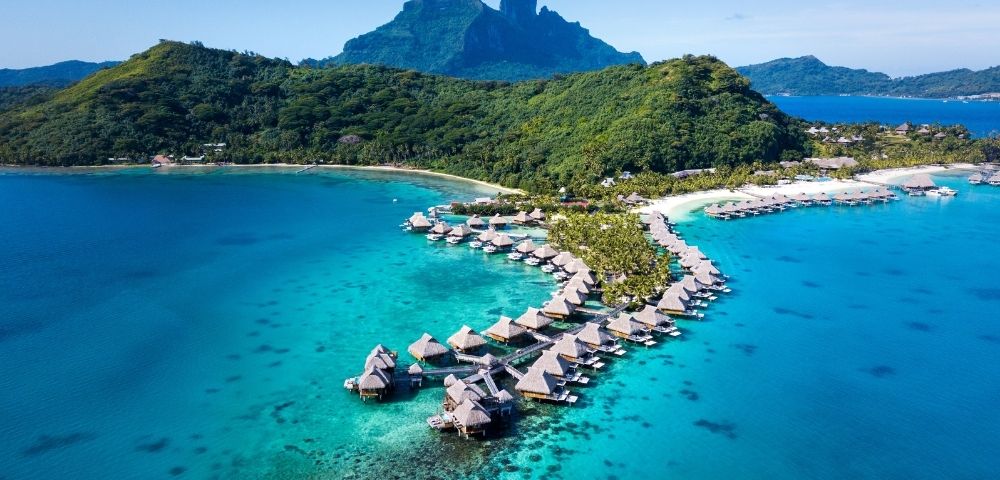 Aerial view of a tropical resort with thatched-roof overwater bungalows extending into a turquoise lagoon, with lush green mountains in the background.