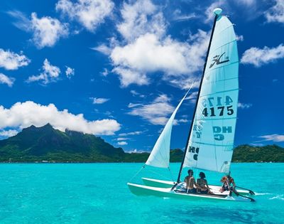 A catamaran sailboat with two people aboard sails across a bright turquoise lagoon, with a lush green, mountainous island under a vivid blue sky dotted with white clouds in the background.