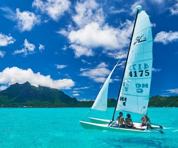 A catamaran sailboat with two people aboard sails across a bright turquoise lagoon, with a lush green, mountainous island under a vivid blue sky dotted with white clouds in the background.