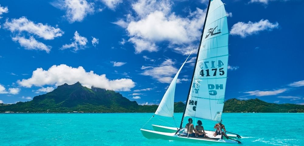 A catamaran sailboat with two people aboard sails across a bright turquoise lagoon, with a lush green, mountainous island under a vivid blue sky dotted with white clouds in the background.