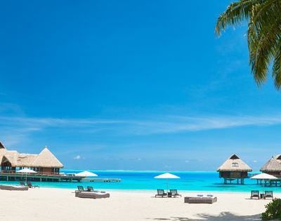 Overwater bungalows with thatched roofs stand on stilts in turquoise water, with a white sandy beach, lounge chairs, and palm trees in the foreground under a bright blue sky.