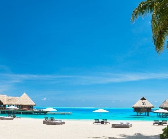 Overwater bungalows with thatched roofs stand on stilts in turquoise water, with a white sandy beach, lounge chairs, and palm trees in the foreground under a bright blue sky.
