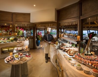 A chef in a white uniform and hat stands behind a lavish buffet filled with seafood, sushi, salads, and an assortment of colorful desserts in a tropical setting with thatched roofs and ocean views in the background.