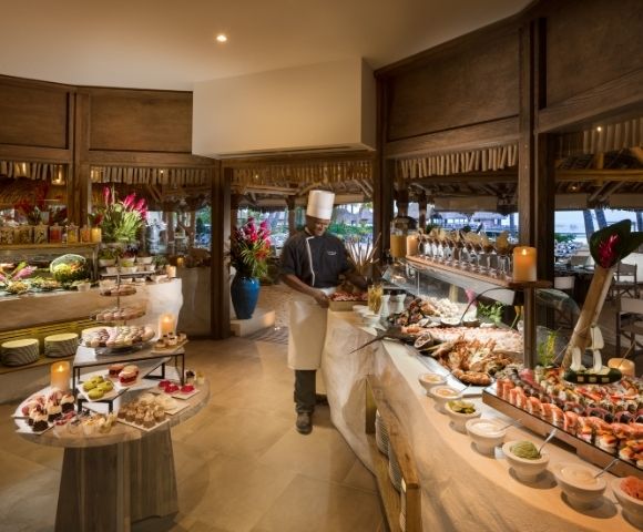 A chef in a white uniform and hat stands behind a lavish buffet filled with seafood, sushi, salads, and an assortment of colorful desserts in a tropical setting with thatched roofs and ocean views in the background.