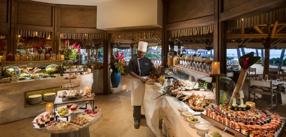 A chef in a white uniform and hat stands behind a lavish buffet filled with seafood, sushi, salads, and an assortment of colorful desserts in a tropical setting with thatched roofs and ocean views in the background.