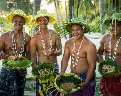 Four men in traditional leaf hats and shell necklaces stand on a beach, each holding a woven leaf basket filled with food.