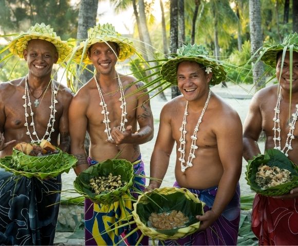 Four men in traditional leaf hats and shell necklaces stand on a beach, each holding a woven leaf basket filled with food.