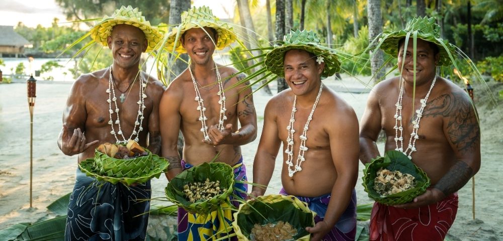 Four men in traditional leaf hats and shell necklaces stand on a beach, each holding a woven leaf basket filled with food.