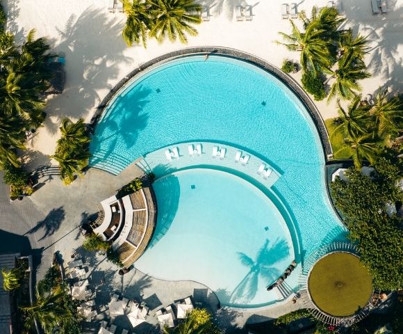 Aerial view of a resort swimming pool shaped like a figure eight, with clear turquoise water, lounge chairs, and surrounding palm trees and sandy beach.