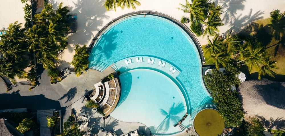Aerial view of a resort swimming pool shaped like a figure eight, with clear turquoise water, lounge chairs, and surrounding palm trees and sandy beach.