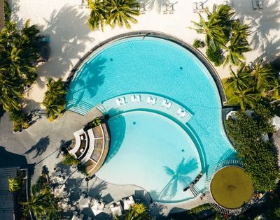 Aerial view of a resort swimming pool shaped like a figure eight, with clear turquoise water, lounge chairs, and surrounding palm trees and sandy beach.
