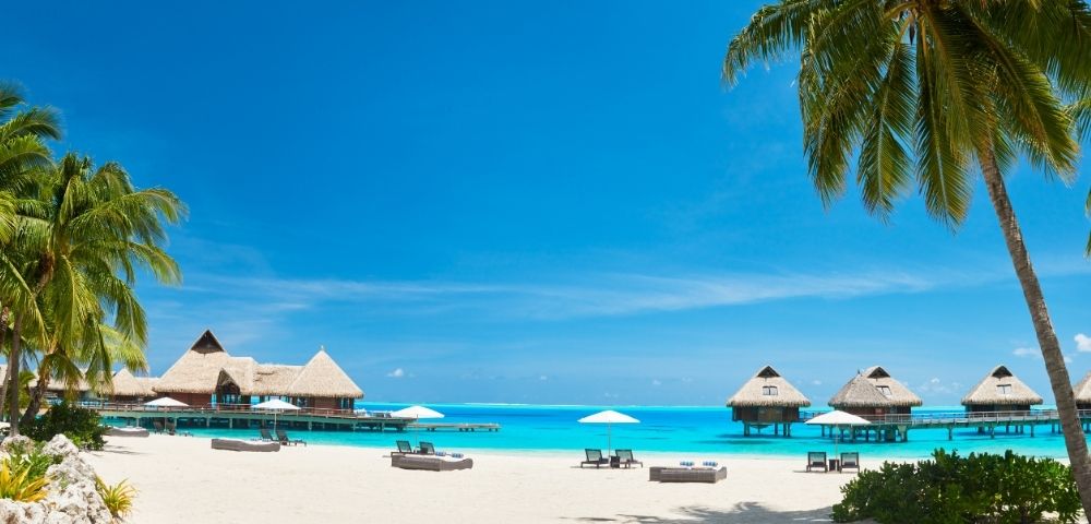 Tropical beach resort with white sand, turquoise lagoon, thatched overwater bungalows, palm trees, lounge chairs, and sunny blue skies.