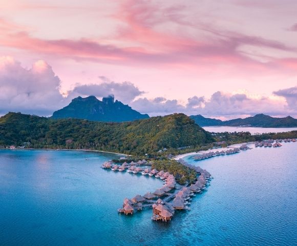 Aerial view of a tropical island resort with turquoise water, curved overwater villas, lush hills, and pink-purple sunset clouds.
