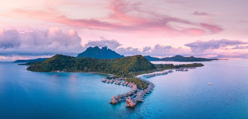 Aerial view of a tropical island resort with turquoise water, curved overwater villas, lush hills, and pink-purple sunset clouds.