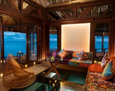 Warm tropical bungalow lounge with rattan chairs, colorful patterned sofa, wood beams, and turquoise ocean views through open windows.