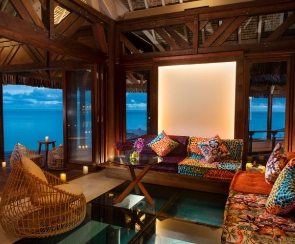 Warm tropical bungalow lounge with rattan chairs, colorful patterned sofa, wood beams, and turquoise ocean views through open windows.