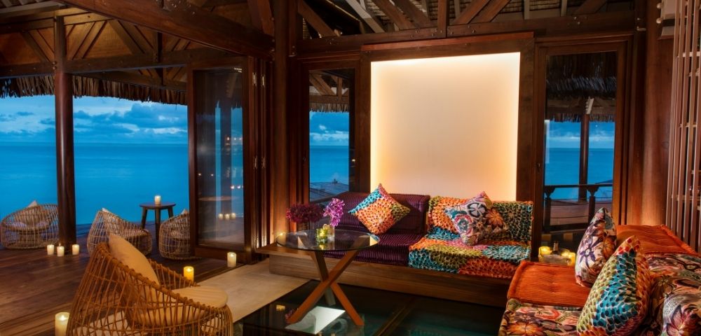 Warm tropical bungalow lounge with rattan chairs, colorful patterned sofa, wood beams, and turquoise ocean views through open windows.