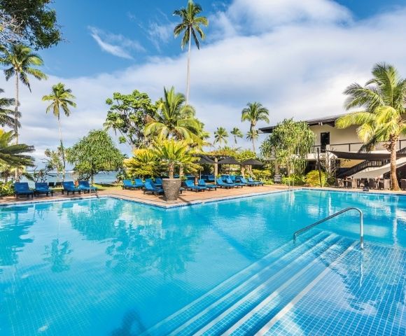 A bright blue swimming pool with steps leading into it, surrounded by lounge chairs and palm trees, with a building in the background.