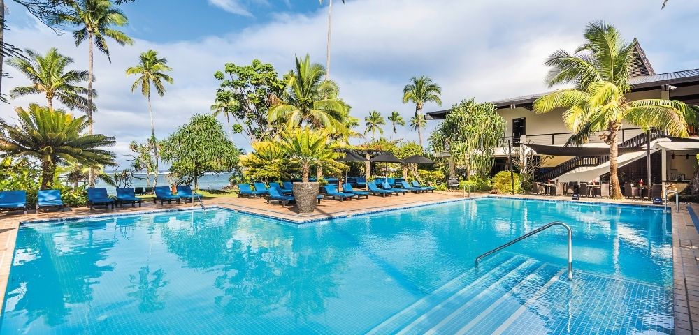 A bright blue swimming pool with steps leading into it, surrounded by lounge chairs and palm trees, with a building in the background.