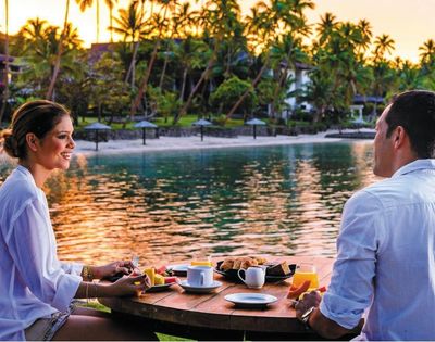 A couple enjoys a scenic breakfast table set with fruit, pastries, coffee, and orange juice on a tropical resort's waterfront.