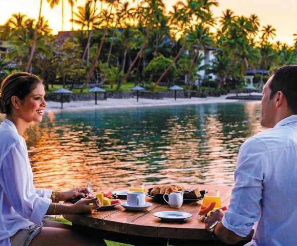 A couple enjoys a scenic breakfast table set with fruit, pastries, coffee, and orange juice on a tropical resort's waterfront.