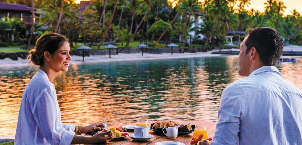 A couple enjoys a scenic breakfast table set with fruit, pastries, coffee, and orange juice on a tropical resort's waterfront.