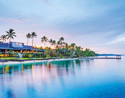 Tropical waterfront resort at dusk, lined with palm trees and glowing lights, reflected in calm blue water beside a long pier.