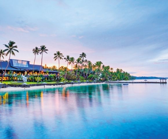 Tropical waterfront resort at dusk, lined with palm trees and glowing lights, reflected in calm blue water beside a long pier.