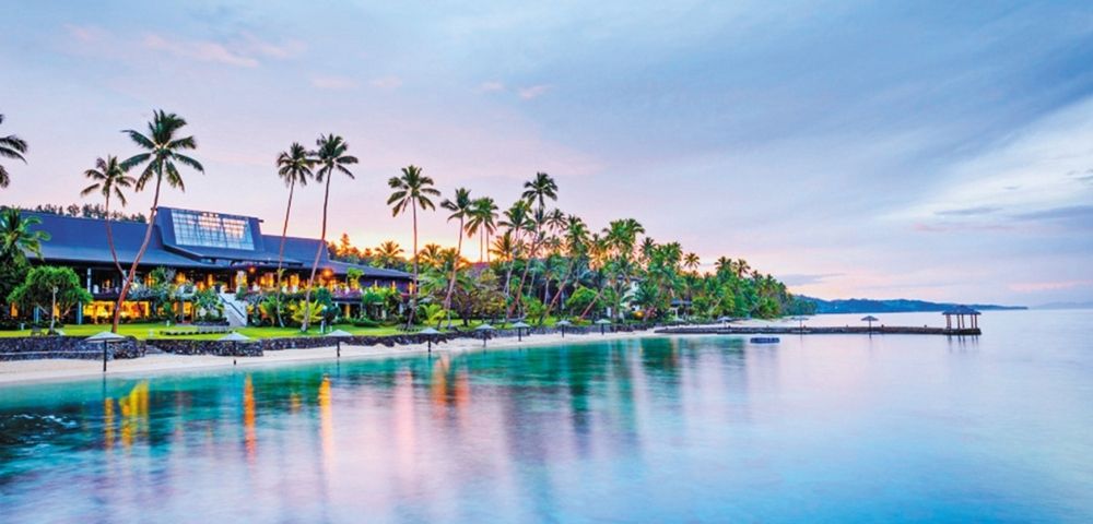 Tropical waterfront resort at dusk, lined with palm trees and glowing lights, reflected in calm blue water beside a long pier.