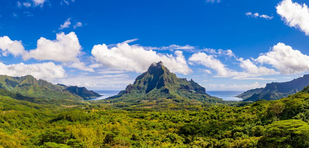 Panoramic view of lush green volcanic mountains surrounding a bay under a vibrant blue sky with scattered white clouds.