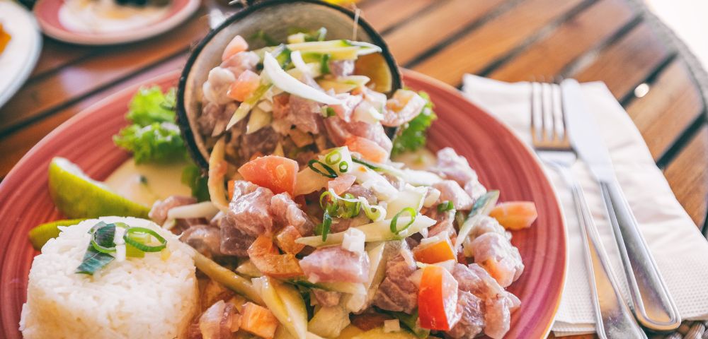 Fresh tuna poke served in a halved coconut shell, with a side of white rice and lime wedge on a red plate at an outdoor table.