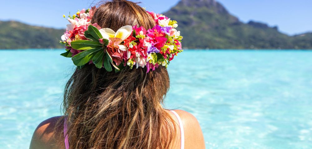 A person in a pink bikini wears a vibrant flower crown as they sit in crystal-clear turquoise water, with a majestic mountain peak visible in the background under a clear blue sky.