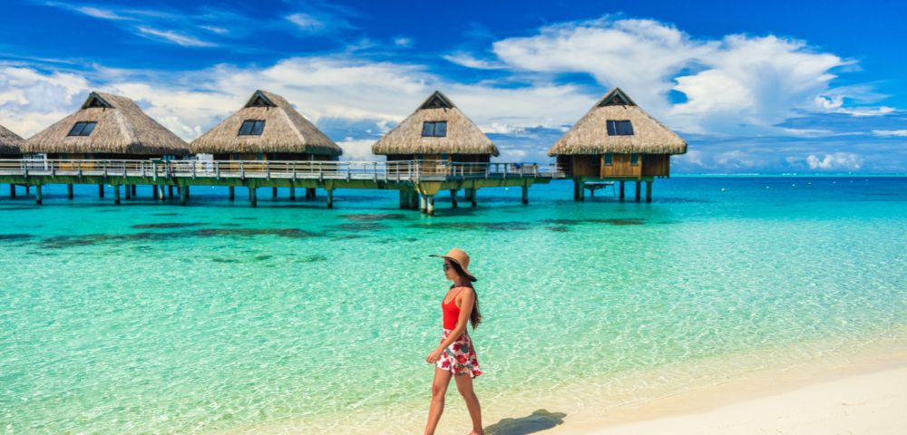 A person walks along a white sand beach on a tropical island, with thatched-roof overwater bungalows in the clear turquoise water under a bright blue sky with white clouds.