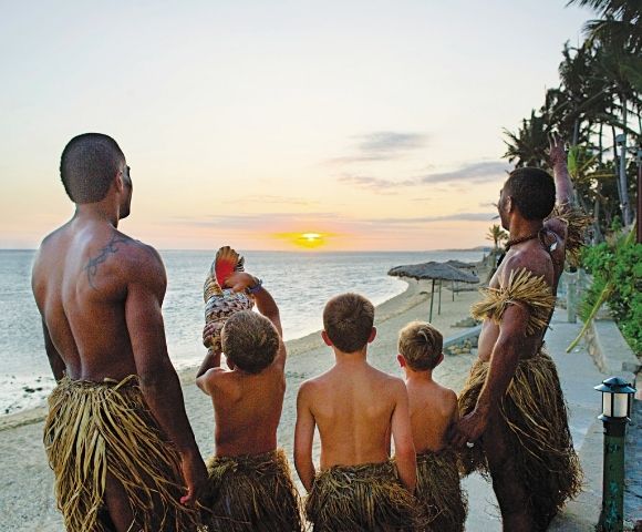Men and boys in traditional grass skirts face the ocean as the sun sets, casting a warm glow over the water and beach.