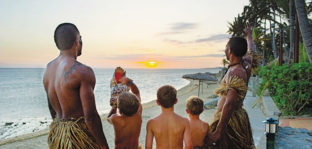 Men and boys in traditional grass skirts face the ocean as the sun sets, casting a warm glow over the water and beach.