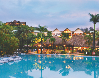 A resort with a large swimming pool in the foreground, illuminated thatched-roof structures in the mid-ground, and buildings on a lush, green hillside in the background, all under a twilight sky.