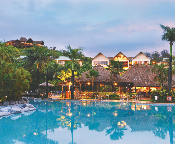 A resort with a large swimming pool in the foreground, illuminated thatched-roof structures in the mid-ground, and buildings on a lush, green hillside in the background, all under a twilight sky.