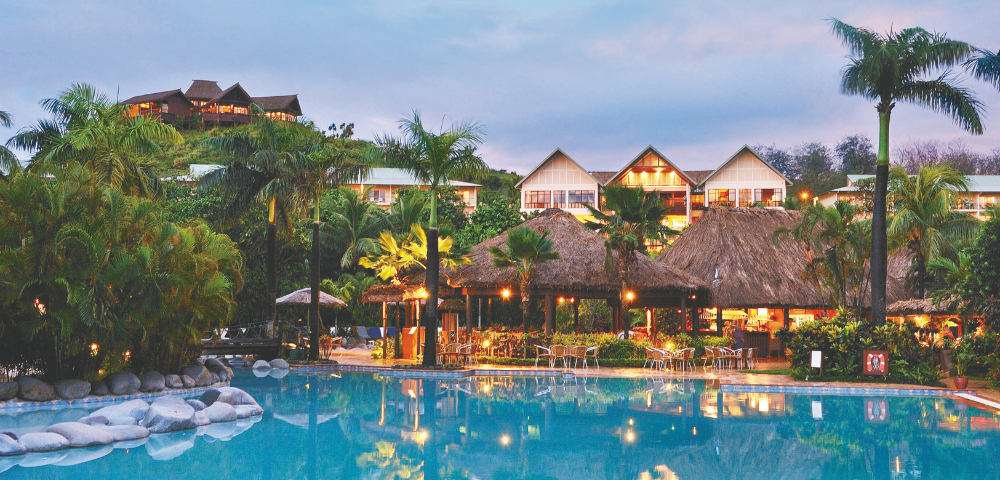 A resort with a large swimming pool in the foreground, illuminated thatched-roof structures in the mid-ground, and buildings on a lush, green hillside in the background, all under a twilight sky.