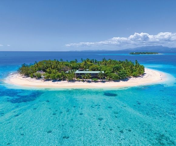 Aerial view of a lush tropical island surrounded by crystal clear turquoise water, featuring a building nestled among palm trees on a white sand beach.