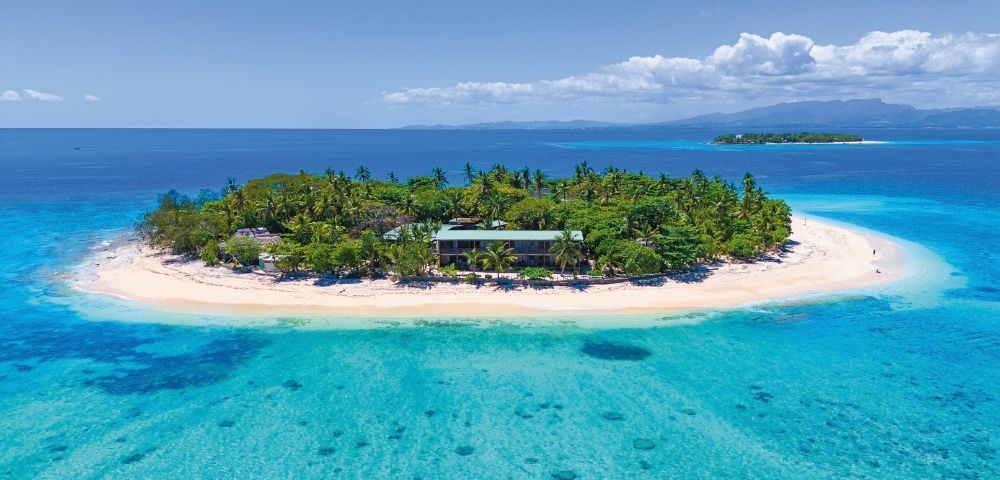 Aerial view of a lush tropical island surrounded by crystal clear turquoise water, featuring a building nestled among palm trees on a white sand beach.