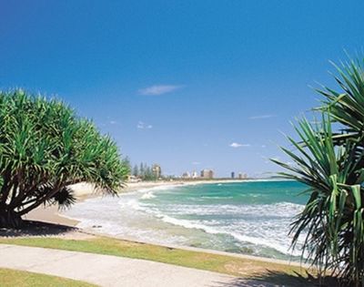 A wide, sandy beach with turquoise ocean waves rolling ashore under a clear blue sky. Lush green trees frame the scene on either side, with a distant city skyline visible along the coast.