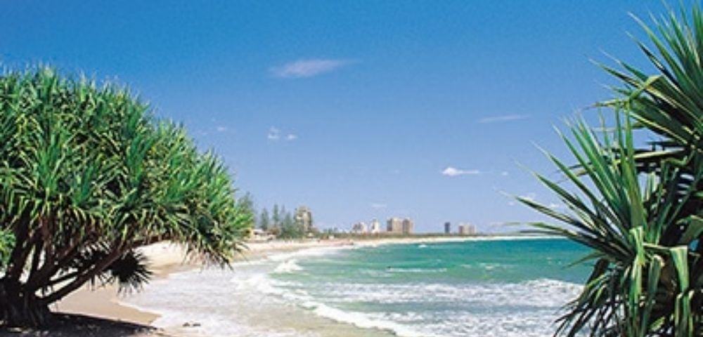A wide, sandy beach with turquoise ocean waves rolling ashore under a clear blue sky. Lush green trees frame the scene on either side, with a distant city skyline visible along the coast.