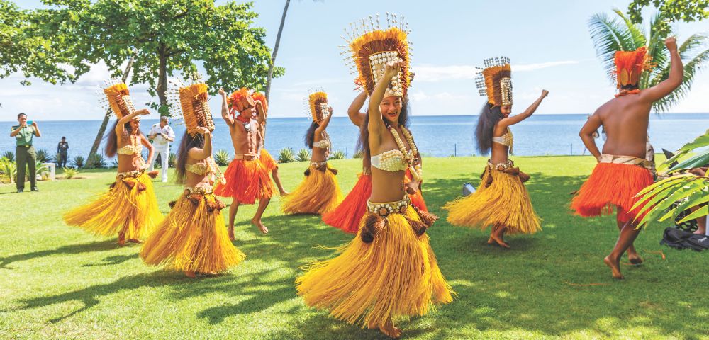 A group of dancers perform a traditional dance on a grassy lawn with the ocean and palm trees in the background. The dancers wear grass skirts in yellow and orange hues, elaborate headdresses, and beaded tops.