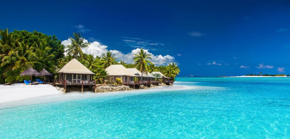 A row of overwater bungalows with thatched roofs sit on stilts above clear turquoise water, surrounded by lush green palm trees and a white sandy beach under a bright blue sky.