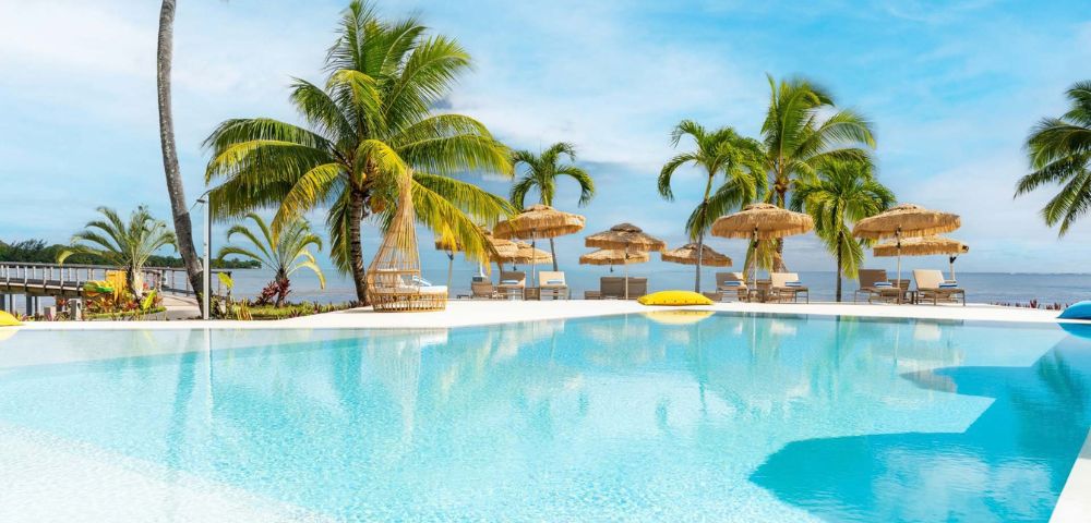 A bright turquoise swimming pool in the foreground reflects the clear blue sky and lush green palm trees. In the background, a row of thatched-roof umbrellas shades lounge chairs on a white deck. The ocean is visible beyond the deck, with a distant shoreline.