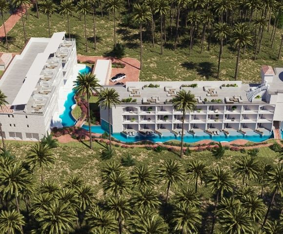 Aerial view of a luxury resort complex surrounded by palm trees, featuring a white modern hotel building with multiple balconies overlooking a winding blue pool and a sandy beach in the background.
