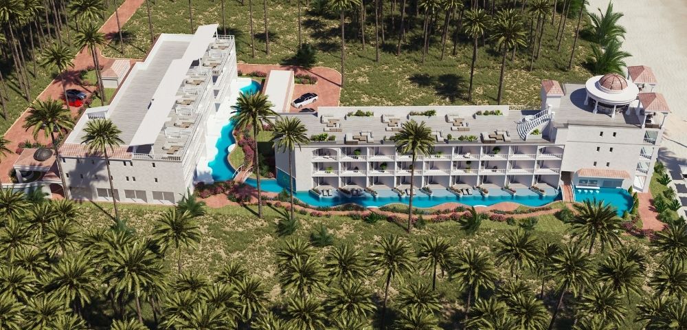 Aerial view of a luxury resort complex surrounded by palm trees, featuring a white modern hotel building with multiple balconies overlooking a winding blue pool and a sandy beach in the background.