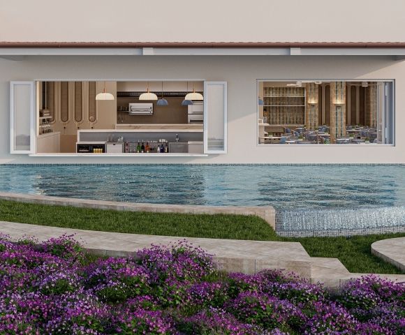A contemporary resort scene with a crescent-shaped swimming pool in the foreground, featuring a cascading waterfall edge. Behind the pool, an open-concept bar and kitchen area is visible through large windows, complete with modern appliances and stocked shelves. To the right, a dining area with tables and chairs is set up under a covered patio, hinting at an adjacent restaurant. Lush green grass and vibrant purple flowers create a serene and inviting atmosphere around the pool.